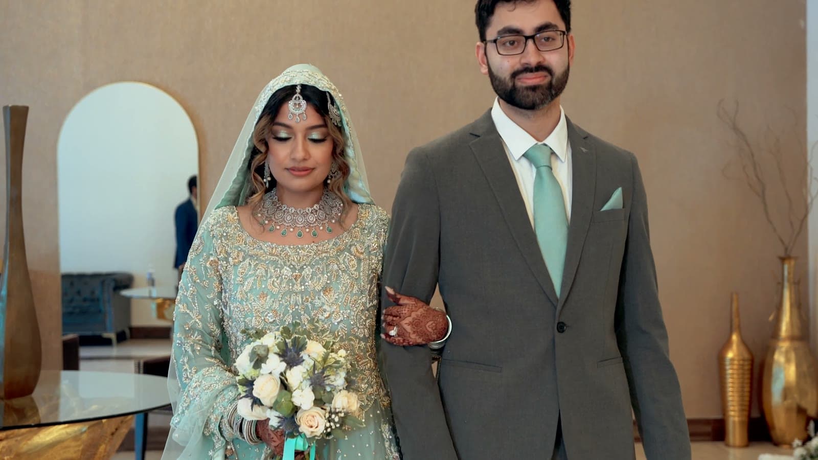 Bride in a mint-and-gold lehenga holding a bouquet beside her groom in a grey suit and mint tie — a still from their nikkah film