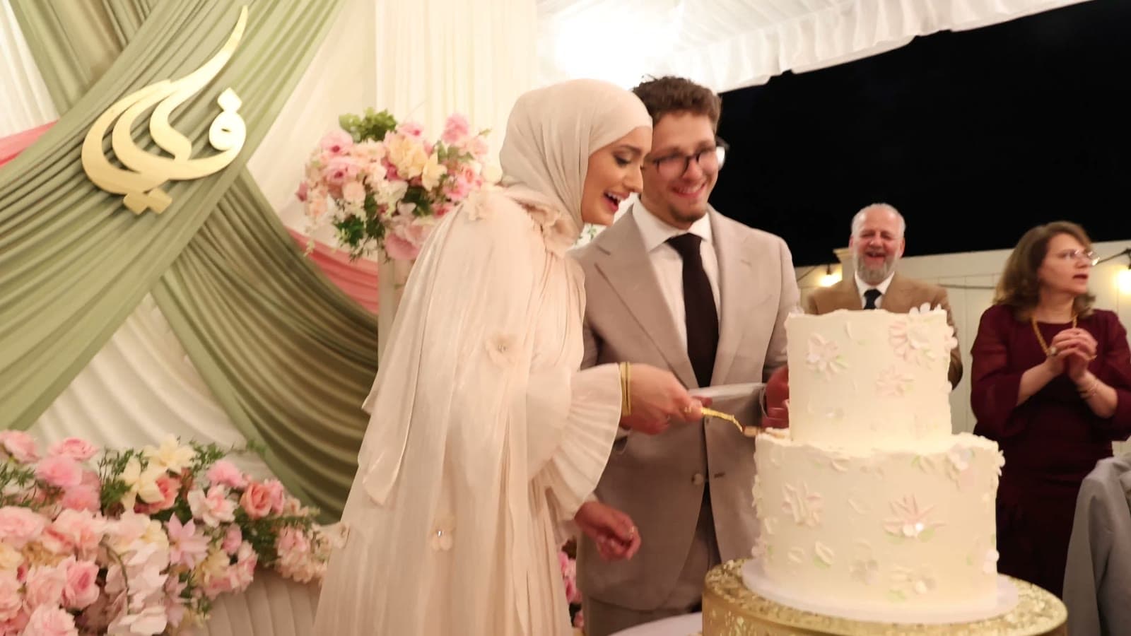 Bride in a cream gown and hijab and groom in a tan suit cutting their flower-topped wedding cake, both laughing — a still from their wedding film