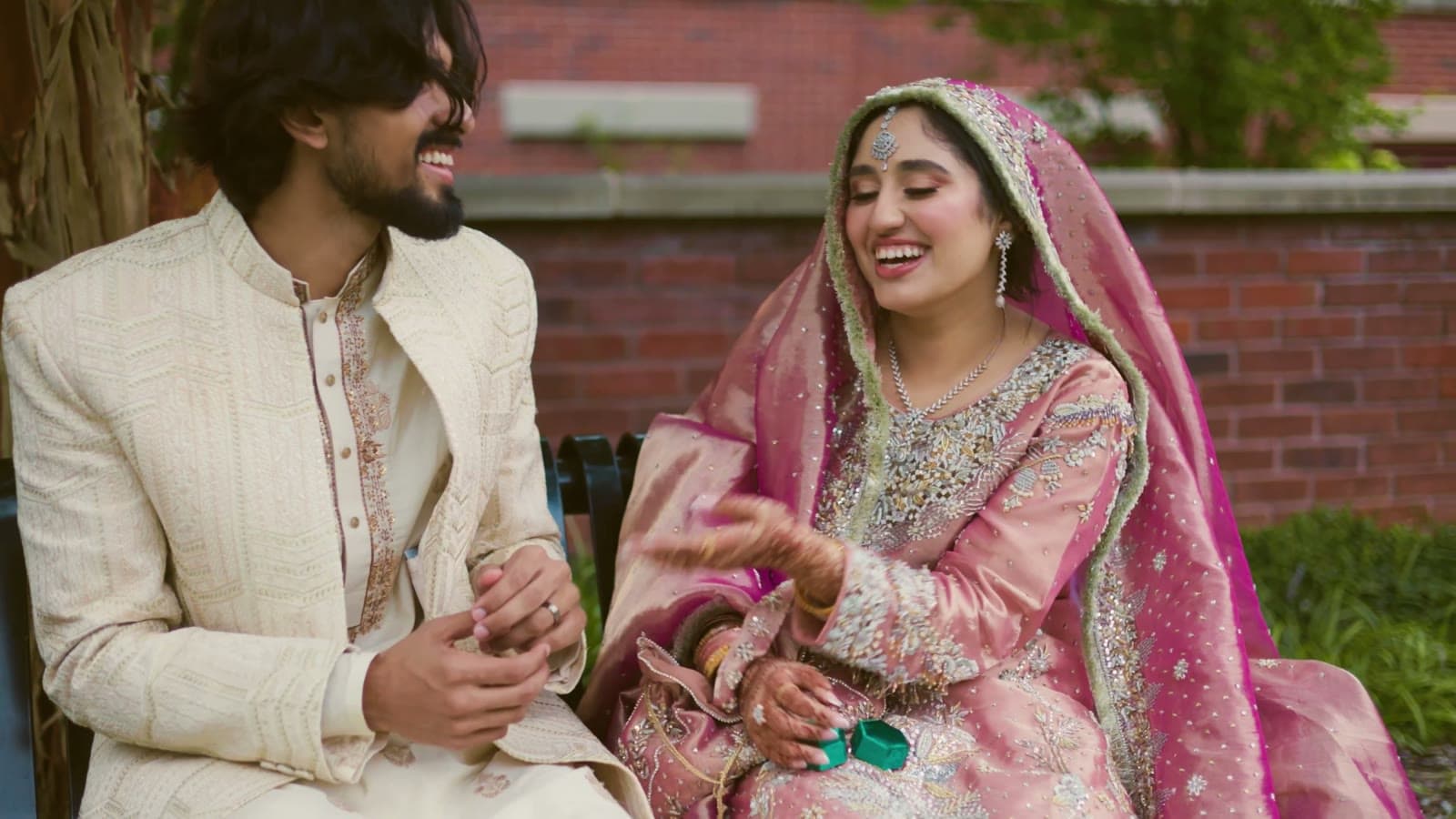 Bride in pink and groom in cream sharing a laugh on a bench, an open ring box in her henna hands — a still from their wedding film
