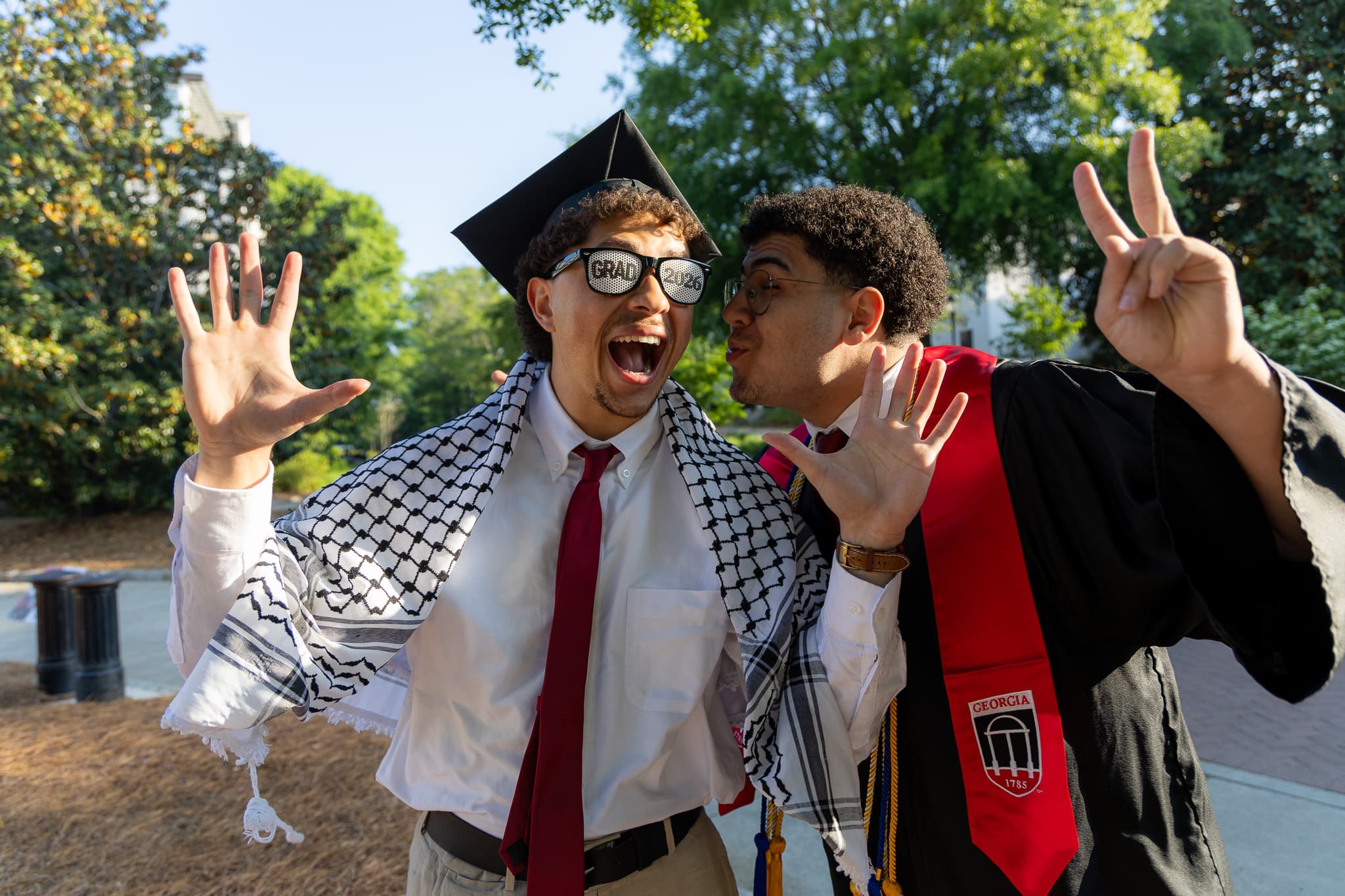 Two UGA graduates in cap, gown, and Georgia stole laughing together on campus at golden hour