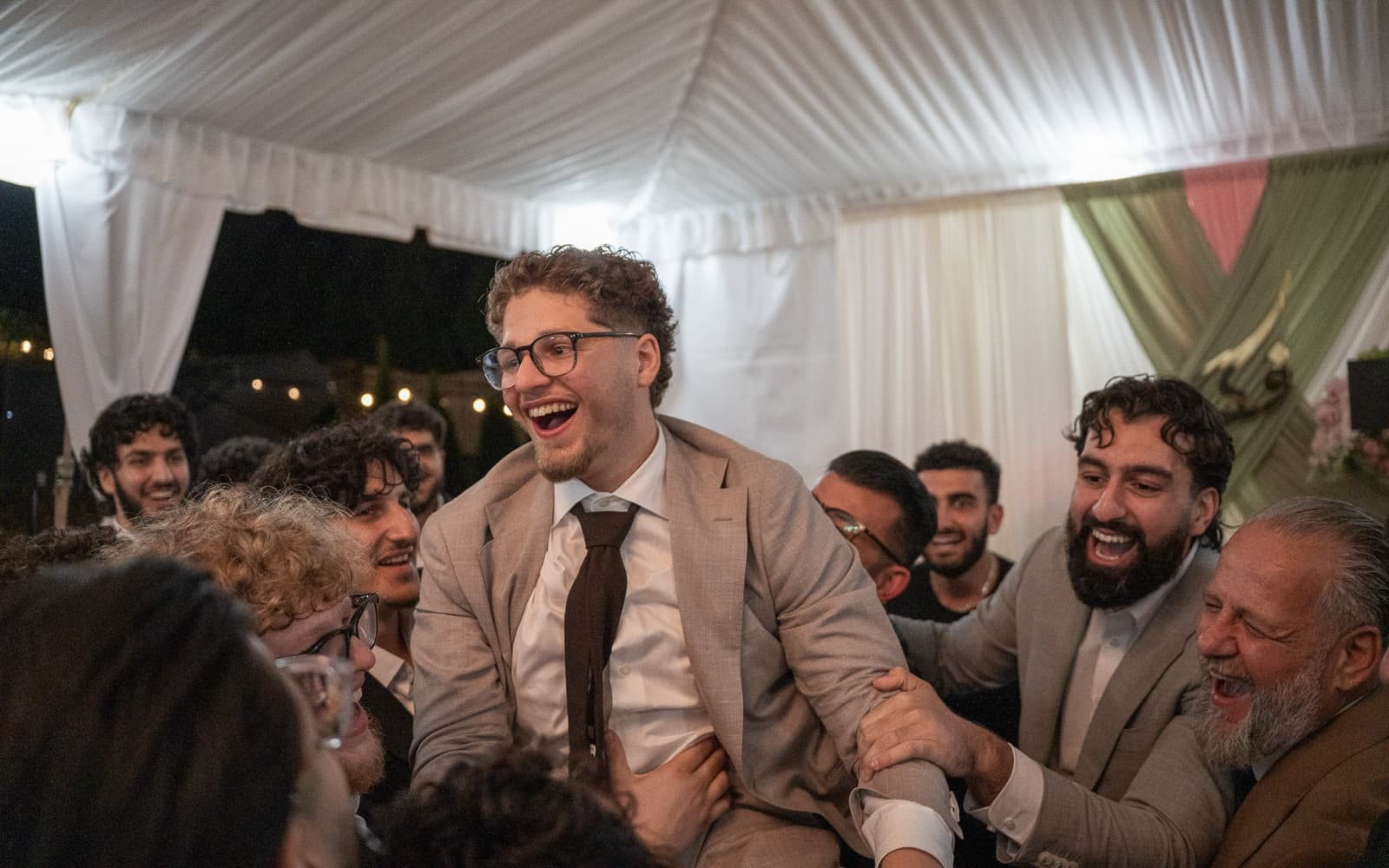 Groom hoisted on shoulders mid-laugh, surrounded by the crowd under the tent at night