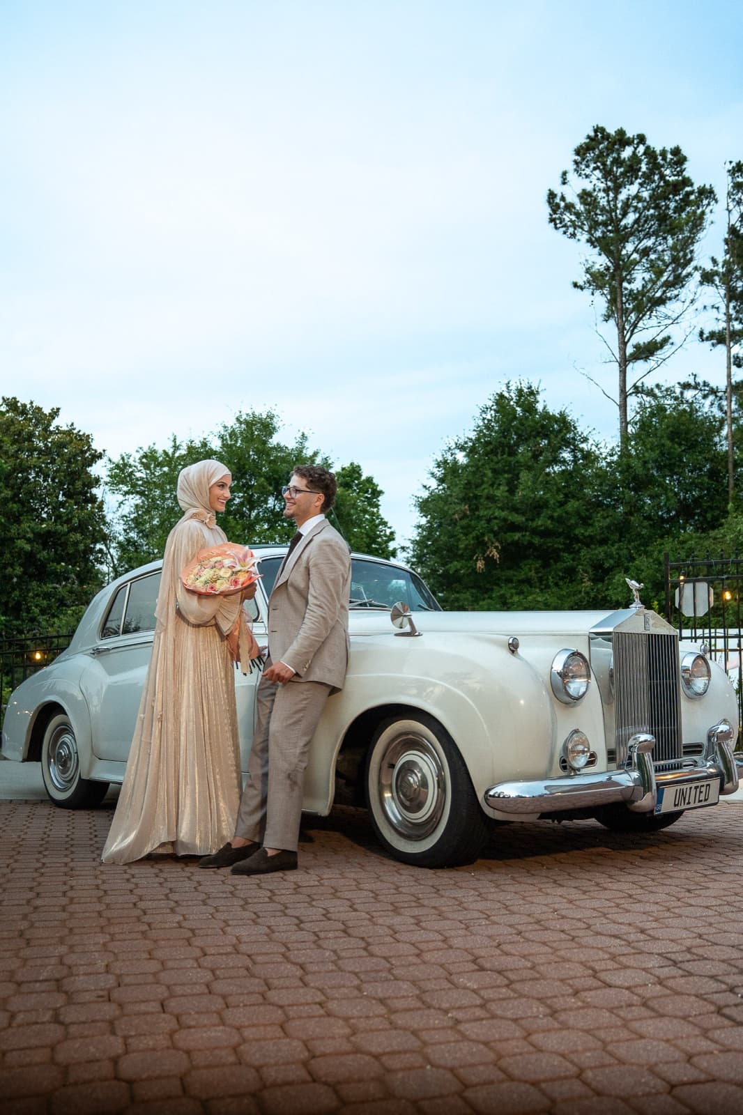 Couple holding a peach bouquet in front of a vintage white Rolls-Royce at dusk