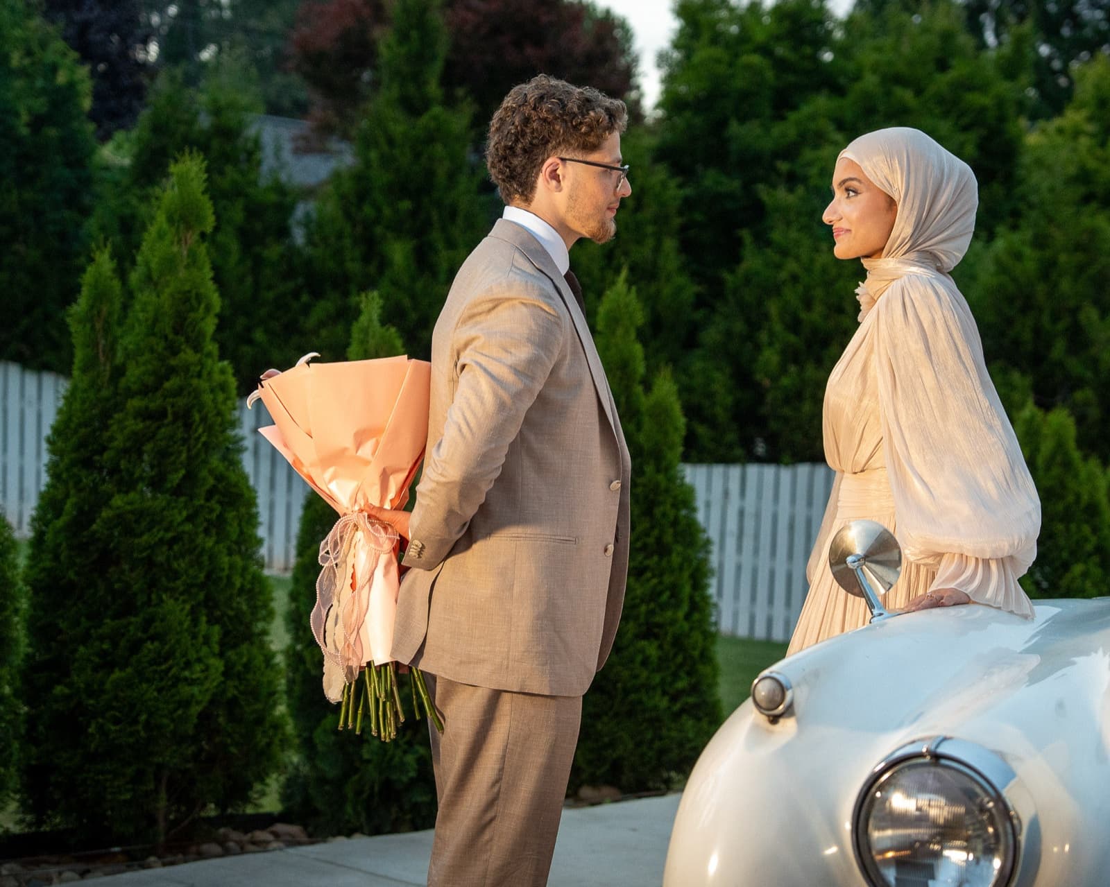 Couple face to face over the fender of a vintage white Rolls-Royce at dusk, bouquet between them