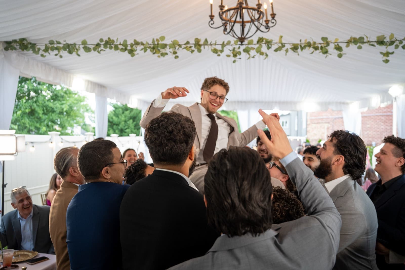 Groom hoisted above the crowd under the tent chandelier, guests reaching up around him