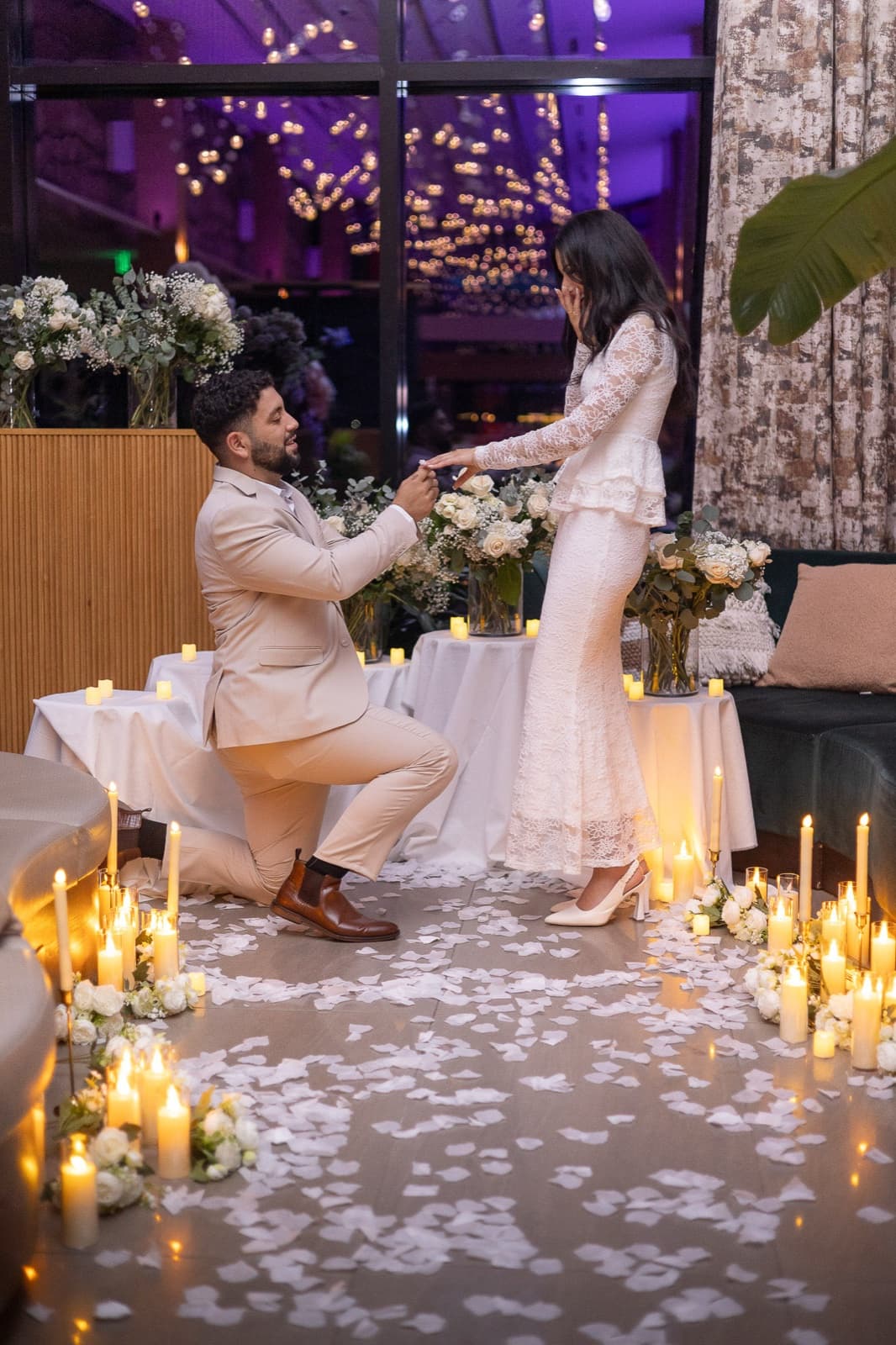 Groom on one knee at a candlelit rooftop proposal, rose petals scattered across the floor