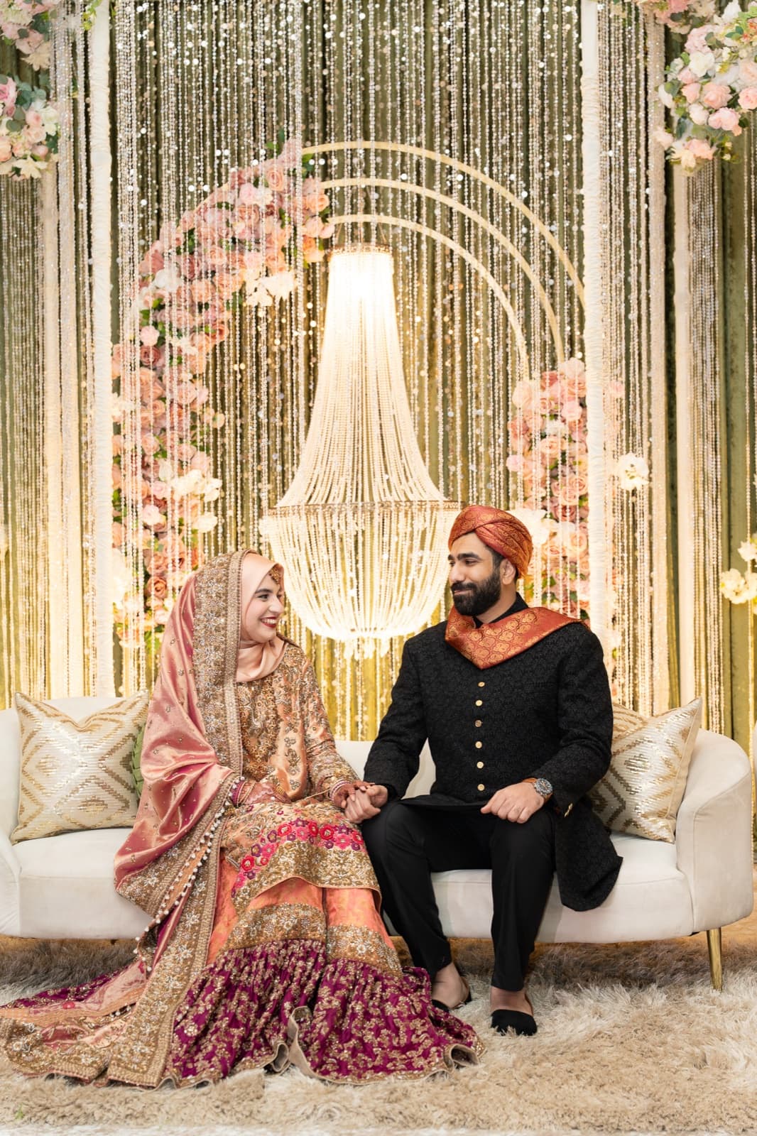 Bride and groom laughing together on a settee beneath a chandelier and cascading light backdrop