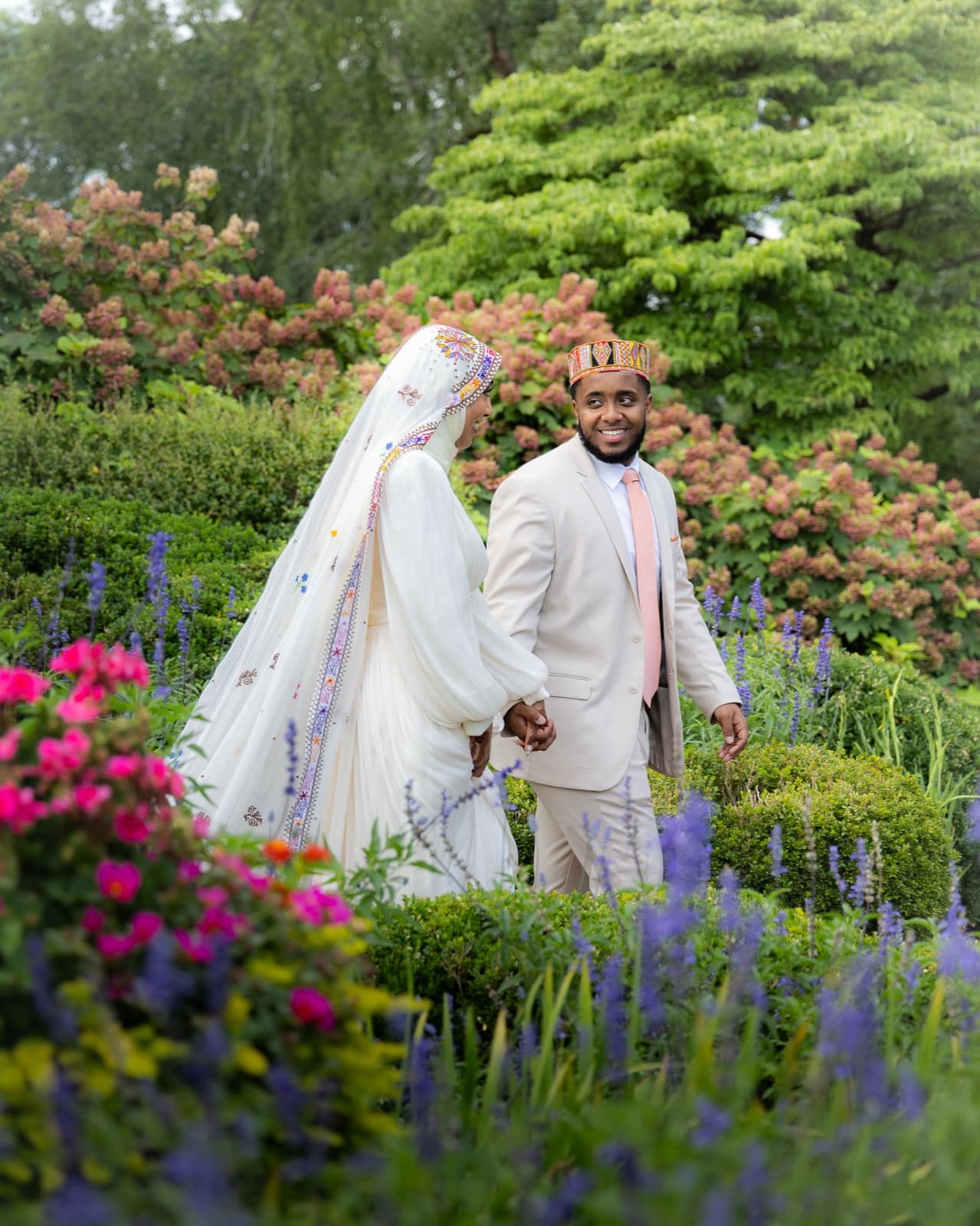 Groom in an embroidered kufi glancing back at his bride as they walk hand in hand through a garden in full bloom