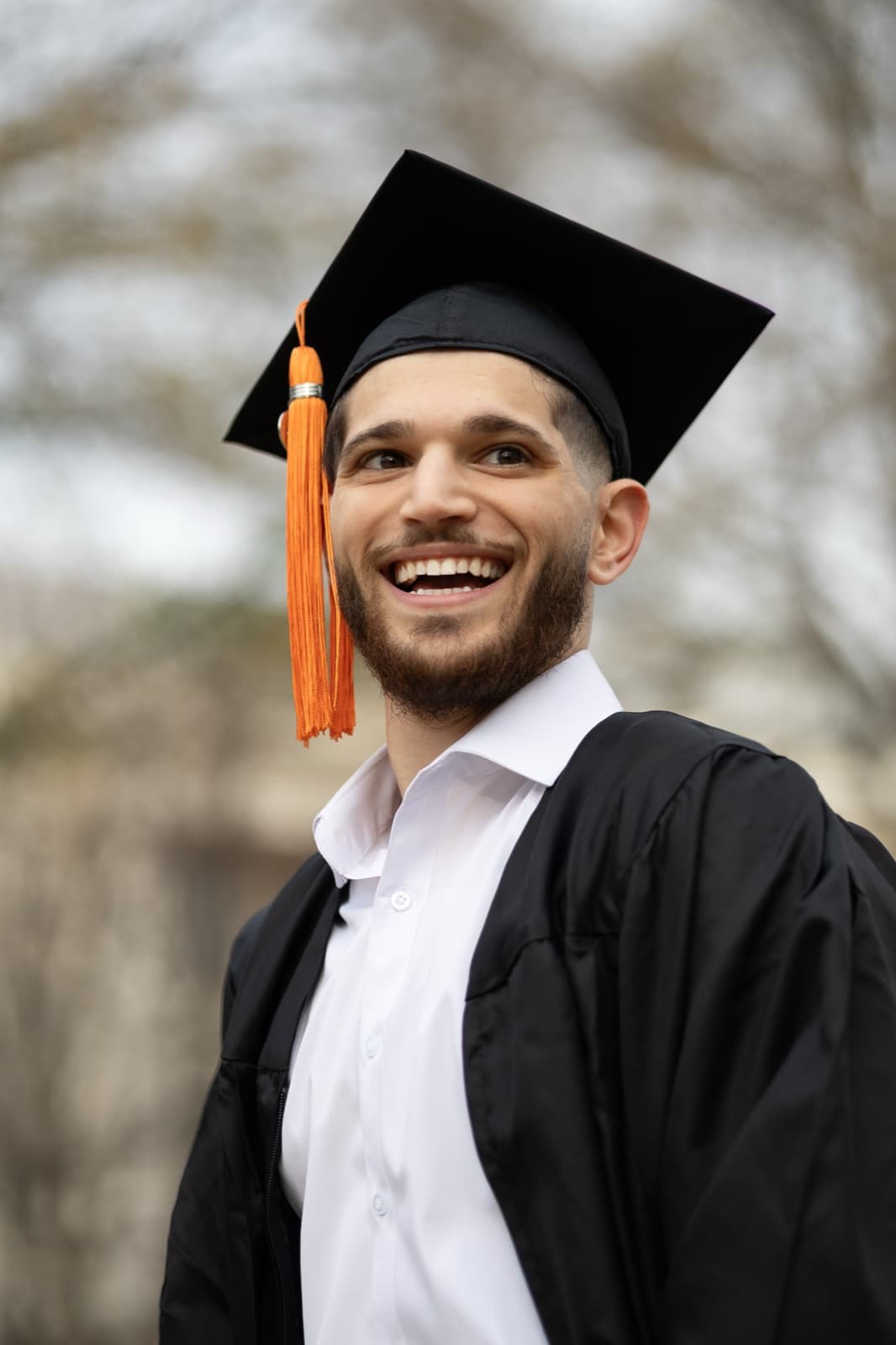 Graduate mid-laugh in cap and gown, orange tassel swinging, on campus