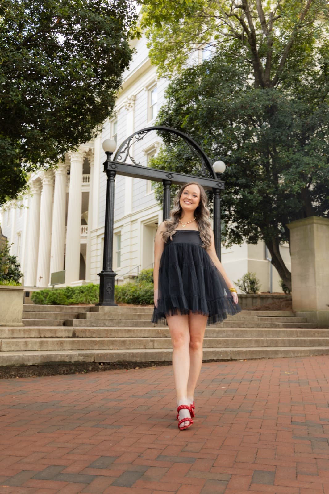 Grad walking in front of the UGA Arch in a black dress and red heels, full length