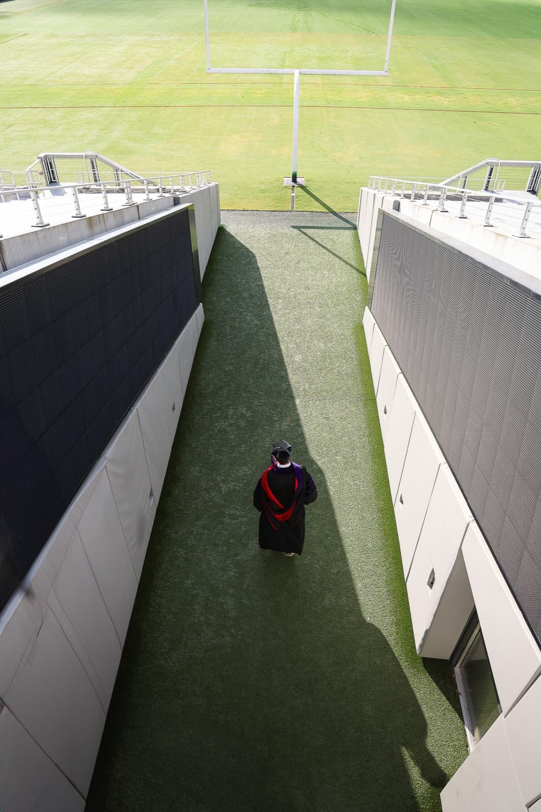 From above: a lone graduate in gown walking the stadium tunnel toward the field