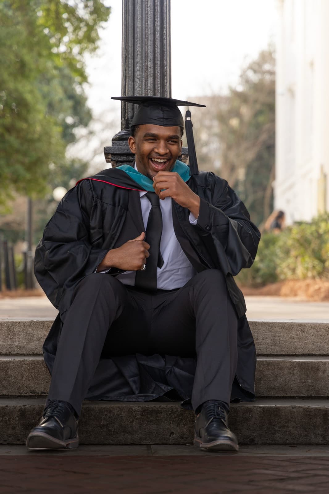 Master's grad cracking up on the steps at the base of a lamppost, hood over his gown