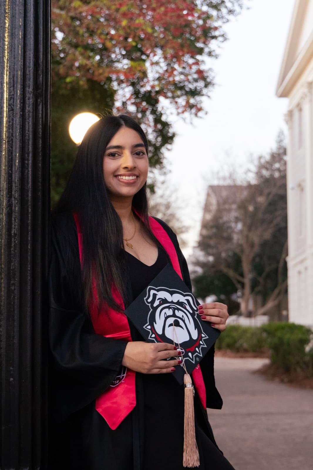 Grad at dusk by an iron gate, holding her Bulldog-painted cap, red UGA stole