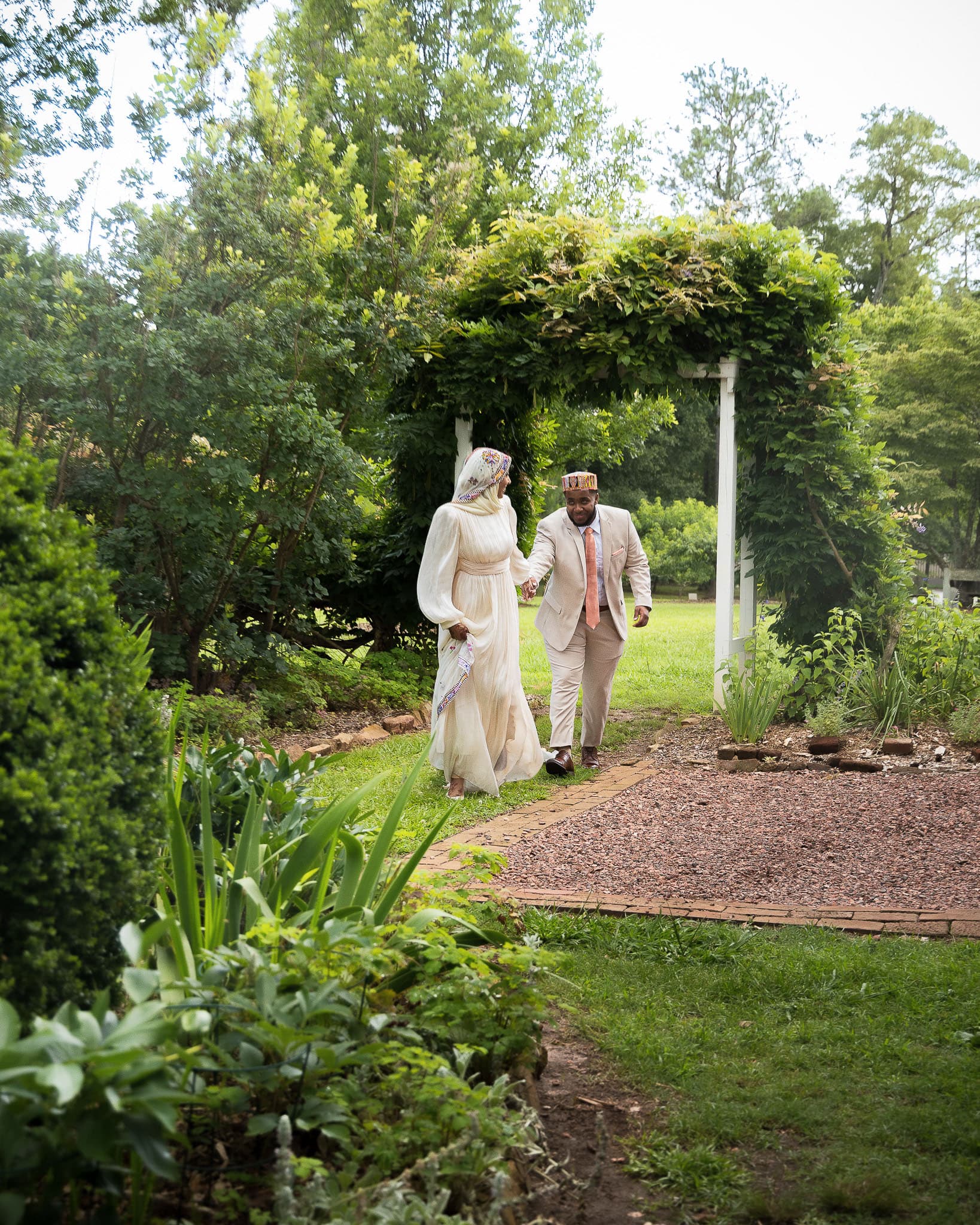 Couple walking hand in hand up a garden path toward a vine-covered arch, her cream gown and veil trailing